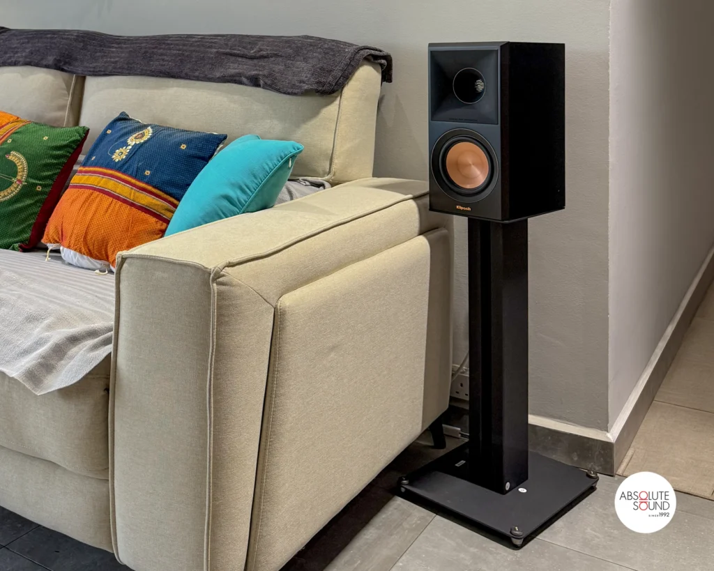 A black Klipsch surround speaker on a floor stand beside a cream sofa with colourful cushions.