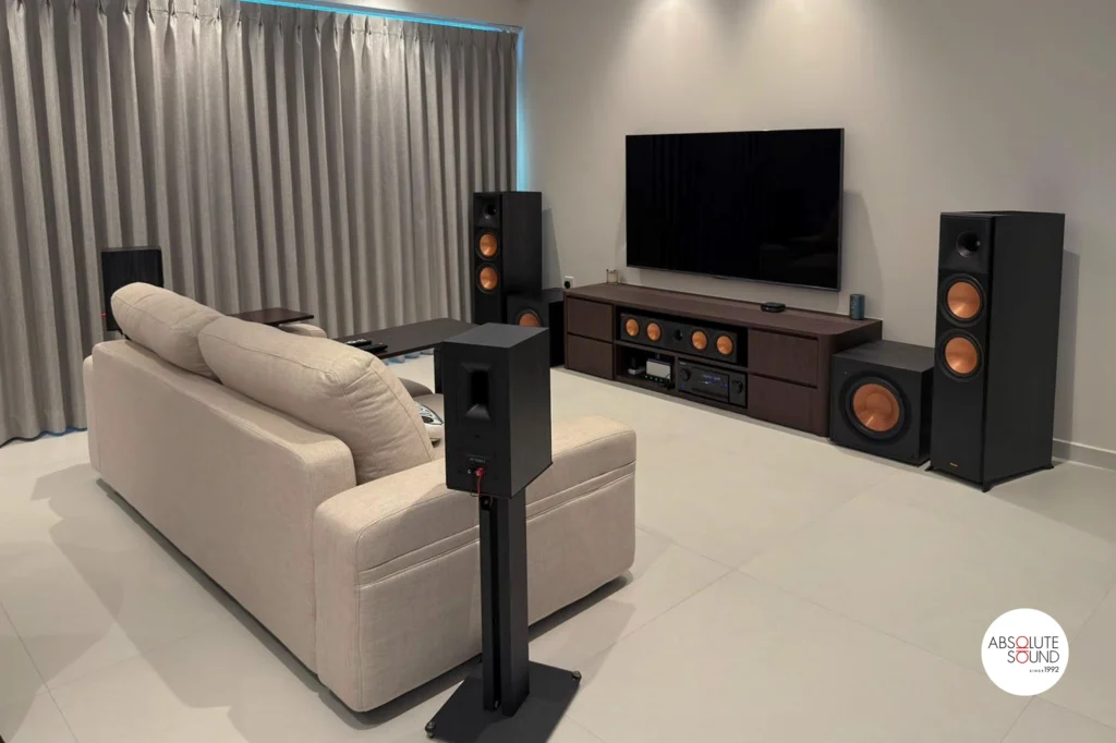 Ebony Klipsch floorstanders and centre speaker along a dark-wood media console below a wall-mounted TV, with a surround on a stand.