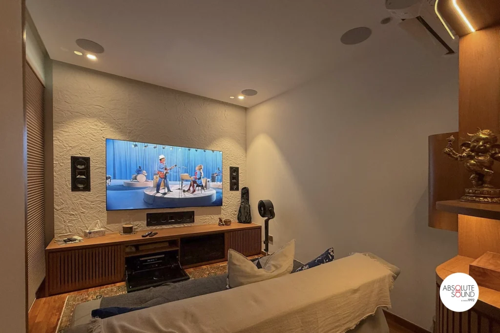 Wall-mounted TV on a white plaster wall, with Klipsch front speakers, grilles off, either side and the centre speaker below.