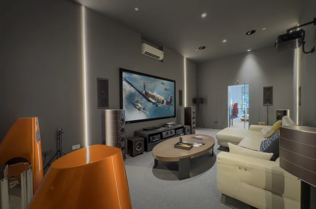 A grey home theatre demonstration room with a large projector screen, floorstanding and in-wall speakers and a cream sofa